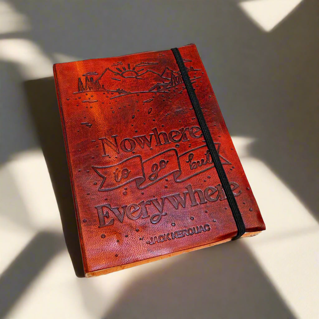 A brown leather journal with an elastic band, featuring an embossed quote and the name 'Jack Kerouac' on the cover, placed on a white wooden surface.