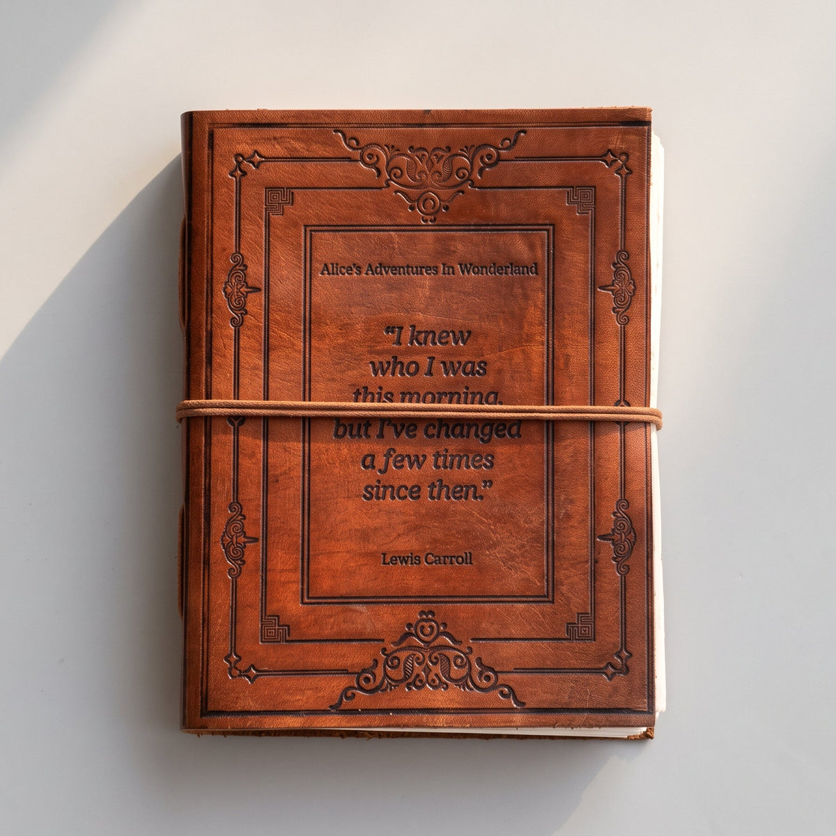 A brown faux-leather notebook or journal with embossed decorative borders and a central quote from Louis Carroll, wrapped with a tan cord, photographed on a light gray wall with soft shadows.
