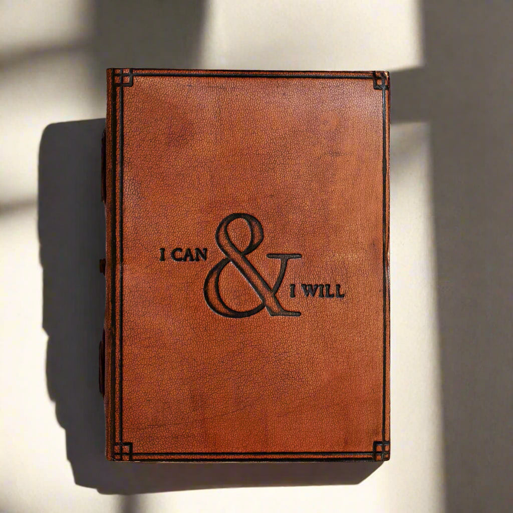 A brown leather journal with the embossed quote 'I Can & I Will' on the cover, placed on a wooden surface.