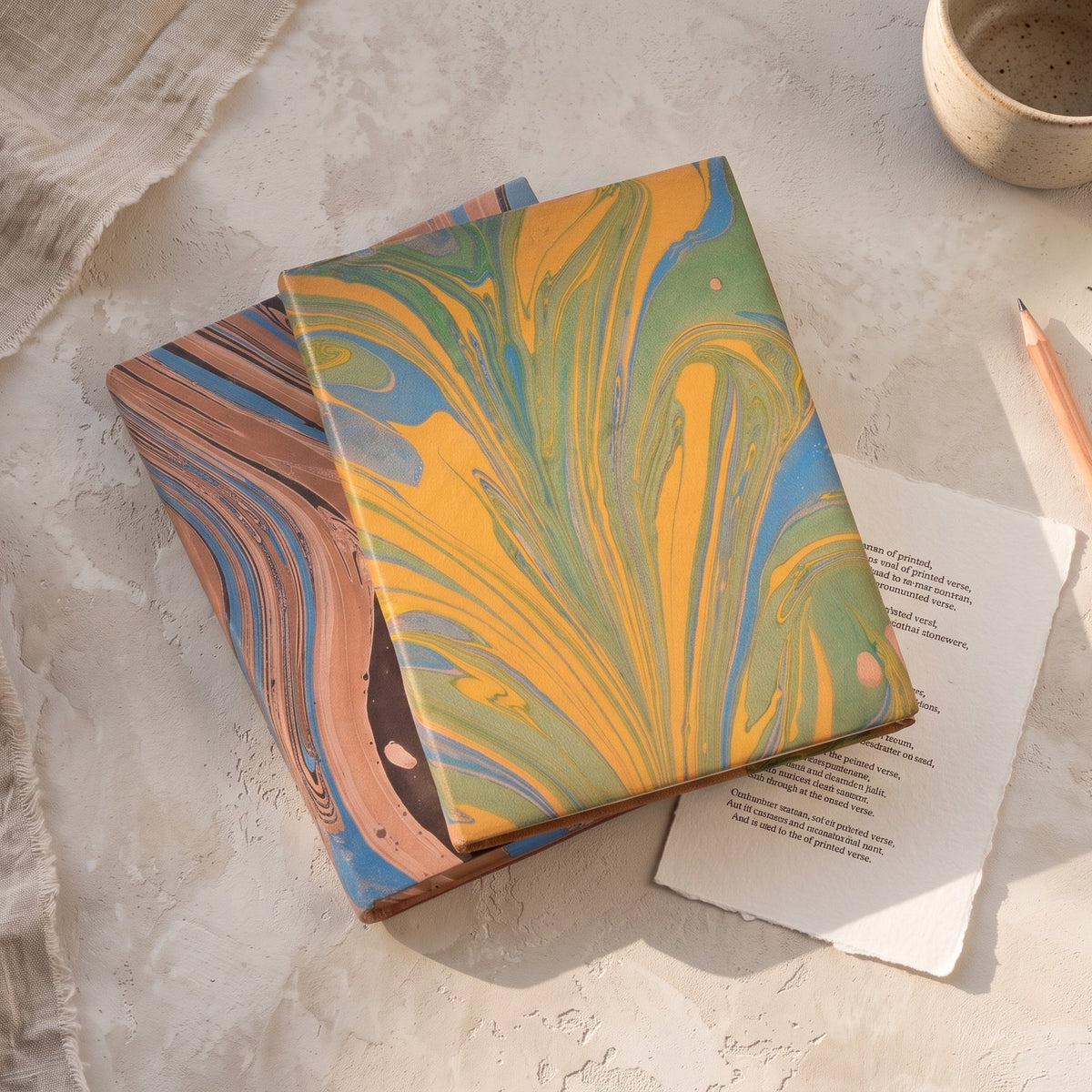 A stack of two decorative notebooks with marbled paper covers in yellow, green, blue, and brown, styled on a light stone surface with a cup, pencil, and paper.