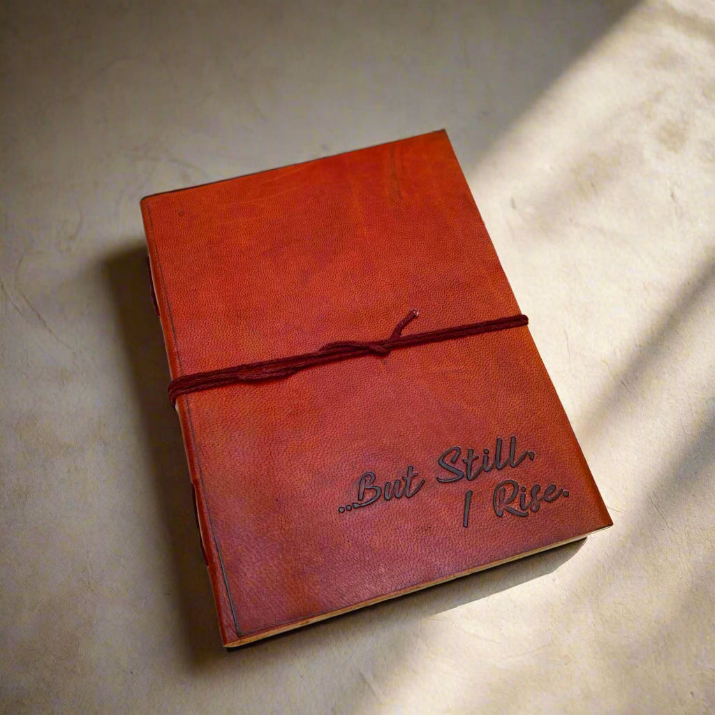 A brown leather journal with the inscription '“But Still I Rise”' embossed on the cover, tied with a red string, placed on a white wooden surface.