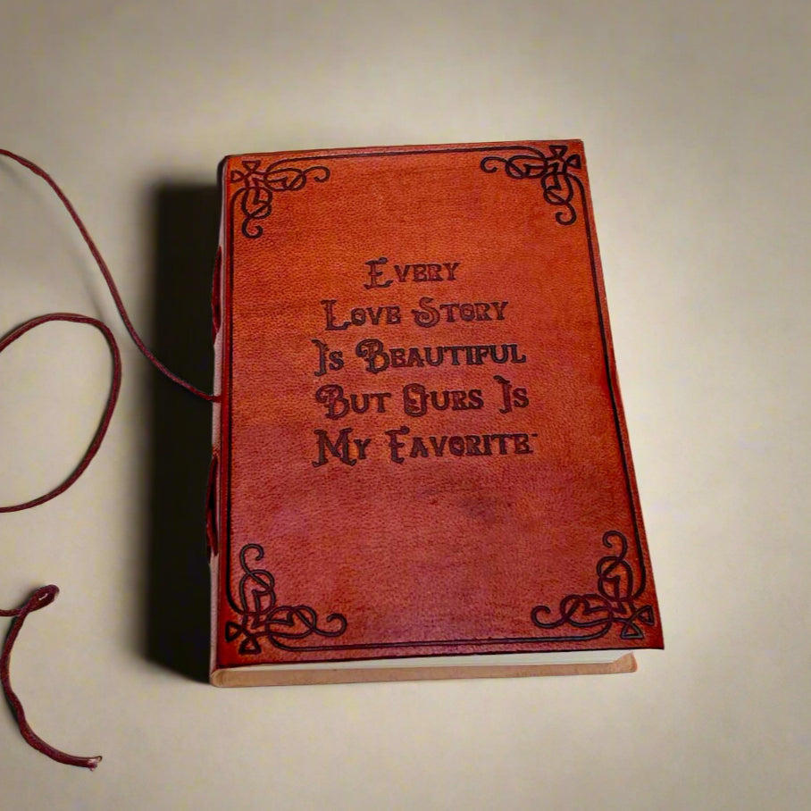 A brown leather journal with a quote embossed on the cover, laying on a white wooden surface. A red string is also visible, likely used for closure.