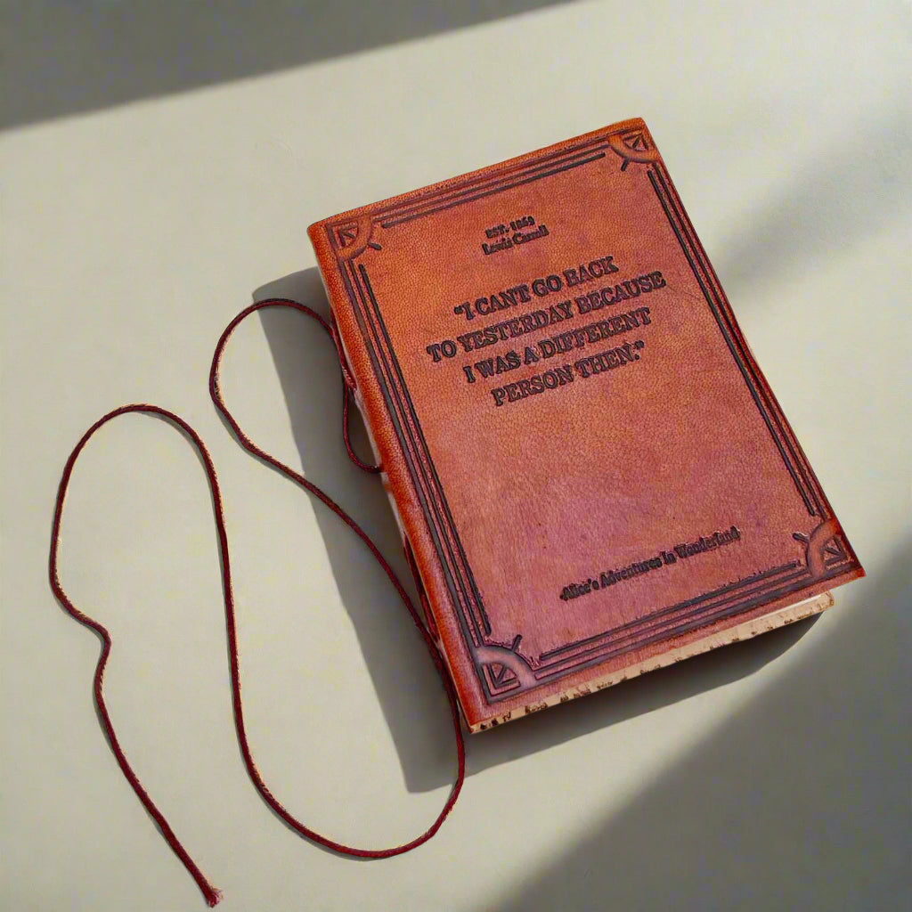 A brown leather journal with a quote from Lewis Carroll's book 'Alice's Adventures in Wonderland' embossed on the cover, placed on a white wooden surface with a red elastic band for closure.