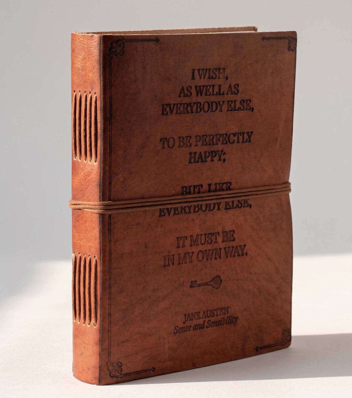 A vintage-style brown leather-bound book or journal standing upright on a light gray background. The cover and spine are brown leather with black embossed text reading, 'I wish, as well as everybody else, to be perfectly happy, but lier everybody else, it must be in my own way.