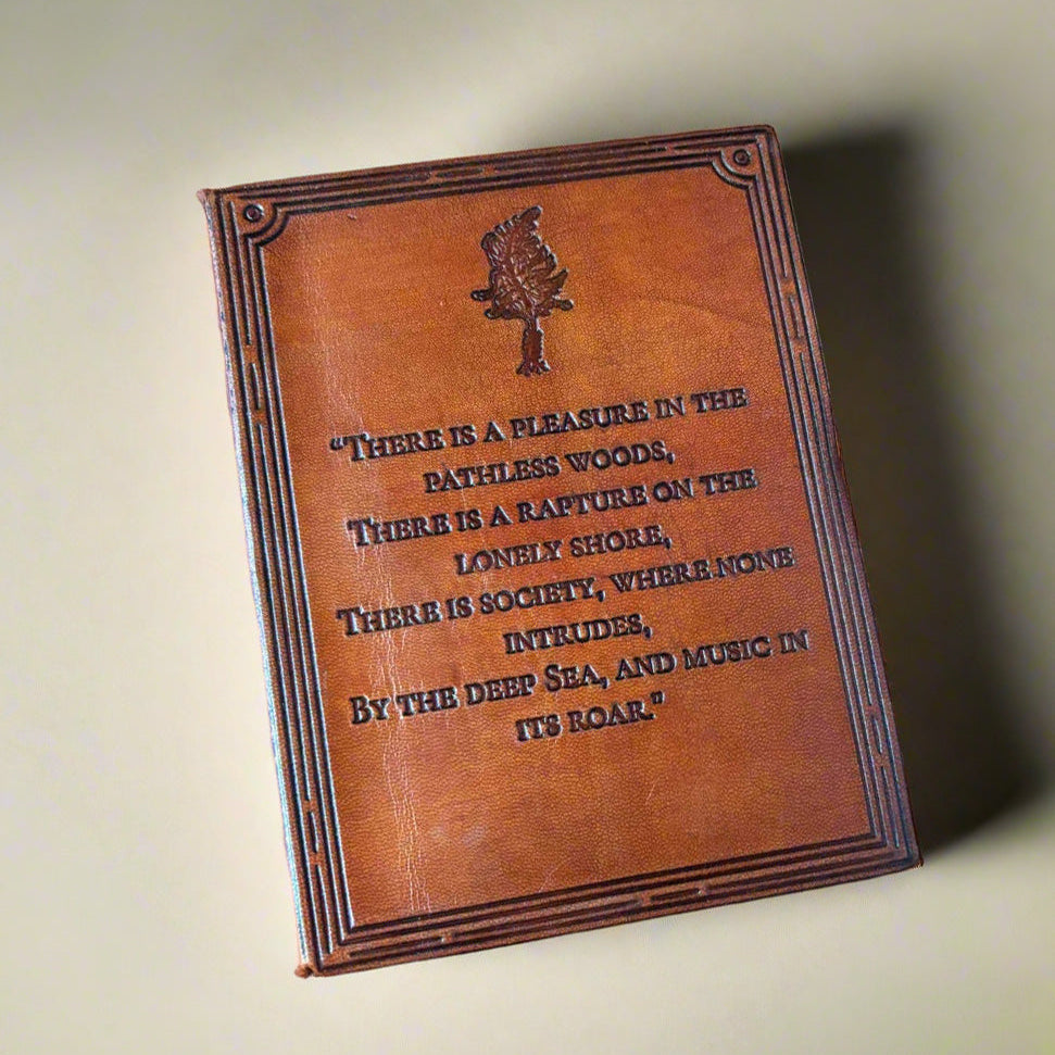 A miniature brown leather-bound book with embossed text and a small tree emblem on the cover, photographed on a neutral beige background.