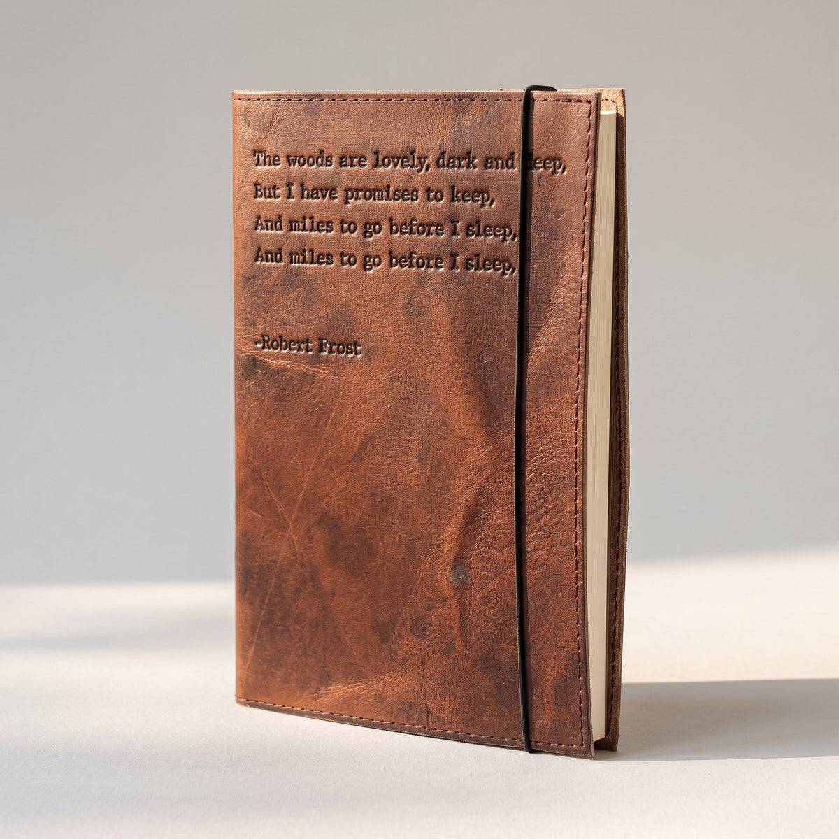 A brown leather journal standing upright on a light gray surface, featuring an embossed quote by Robert Frost on the front cover.