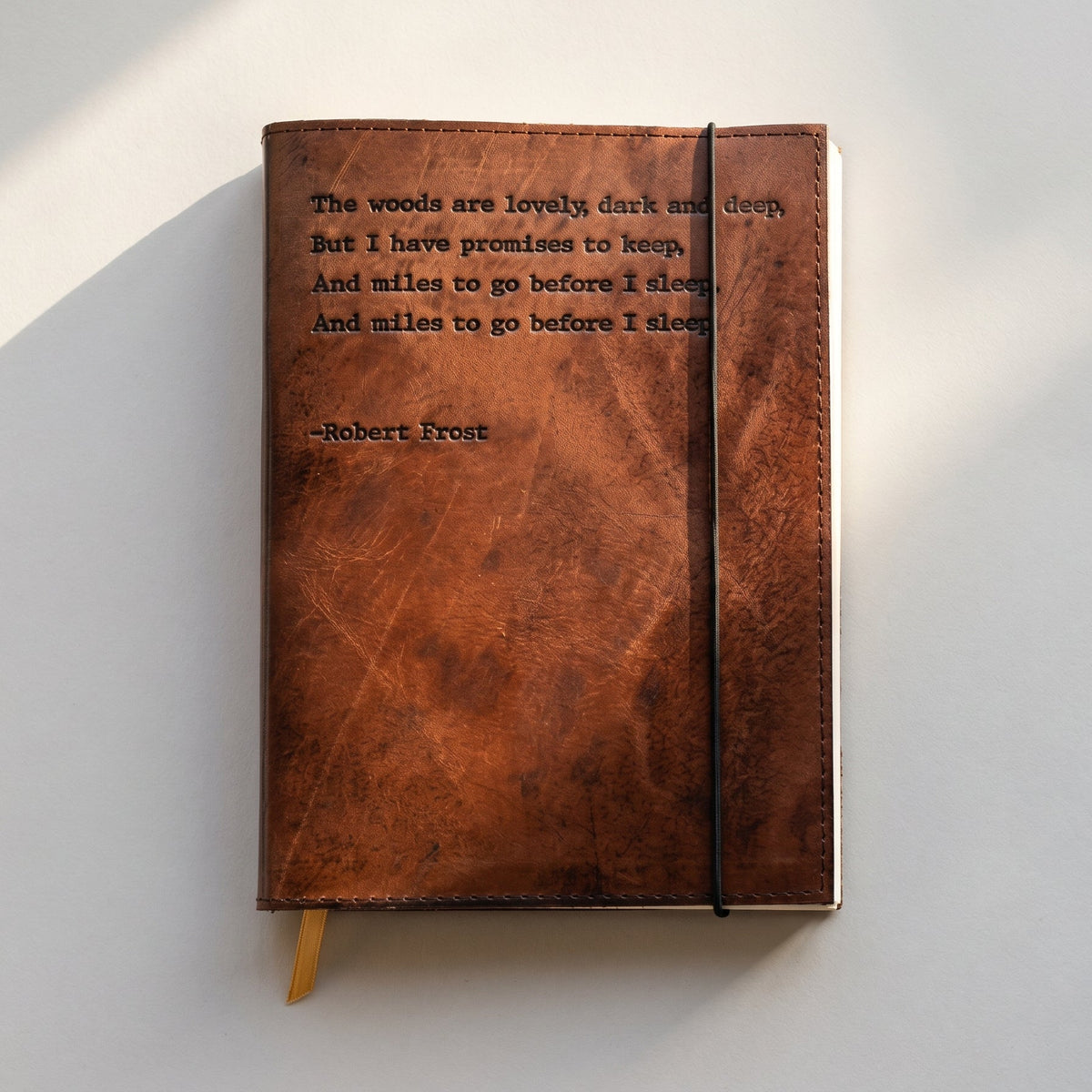 A brown leather-bound journal with an embossed Robert Frost quote on the cover, featuring a black elastic closure and a small gold ribbon bookmark, photographed on a white surface with soft diagonal shadows.