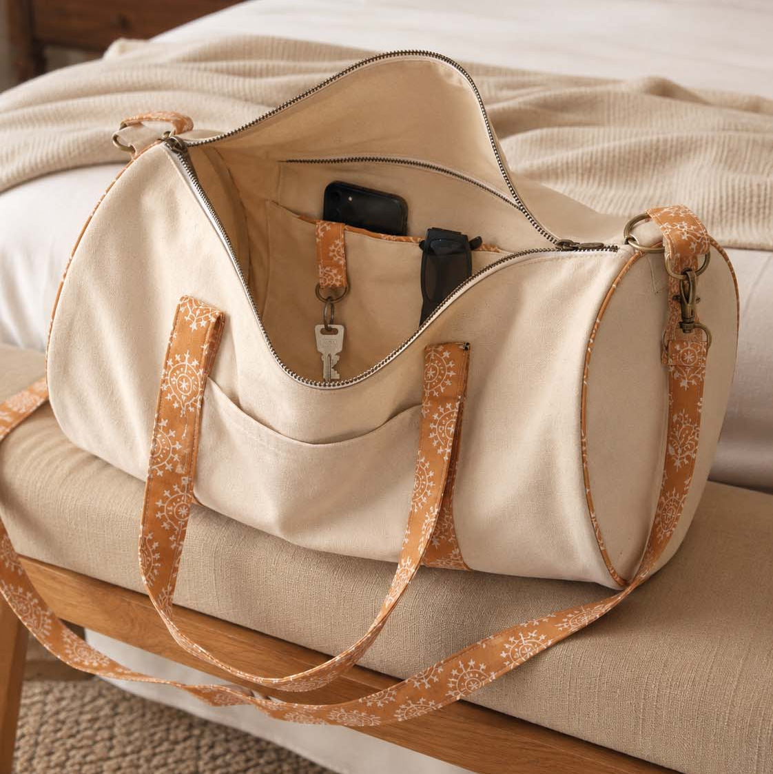 An off-white canvas duffel bag with tan floral-patterned straps and trim, shown open on a bed in a warm, cozy bedroom setting.