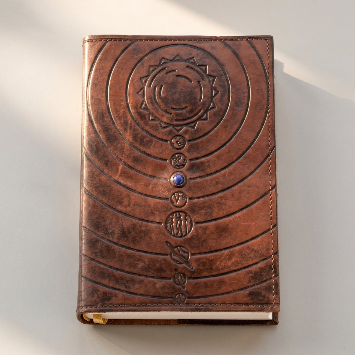 A brown leather journal with embossed solar system and a central blue gem closure, photographed on a light surface with soft shadows.