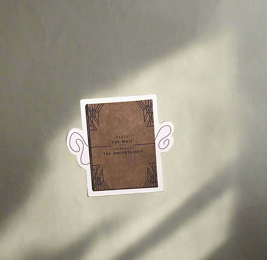 A small rectangular sticker or card with a brown textured center, decorative corner patterns, and centered text reading 'Trust the wait. Embrace the Uncertainty,' shown on a light gray background with soft diagonal shadows.