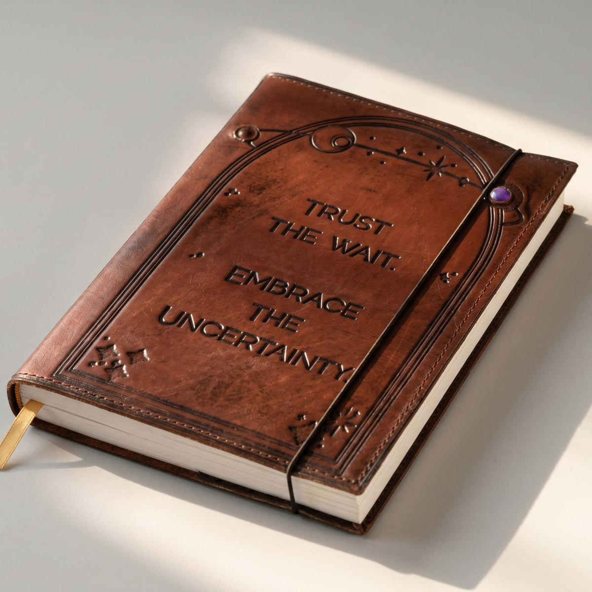 A brown leather-bound journal with embossed gothic-style border details and the words 'TRUST THE WAIT. EMBRACE THE UNCERTAINTY.