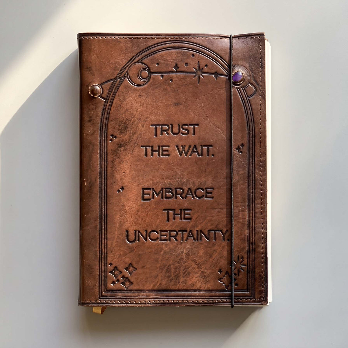 A brown leather-bound journal with embossed decorative border details and the words 'TRUST THE WAIT. EMBRACE THE UNCERTAINTY.
