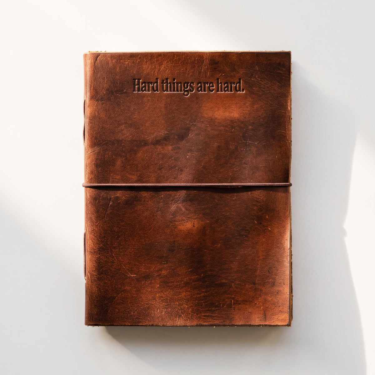 Front view of a brown leather journal with the debossed words “Hard things are hard.” and a string tie across the cover
