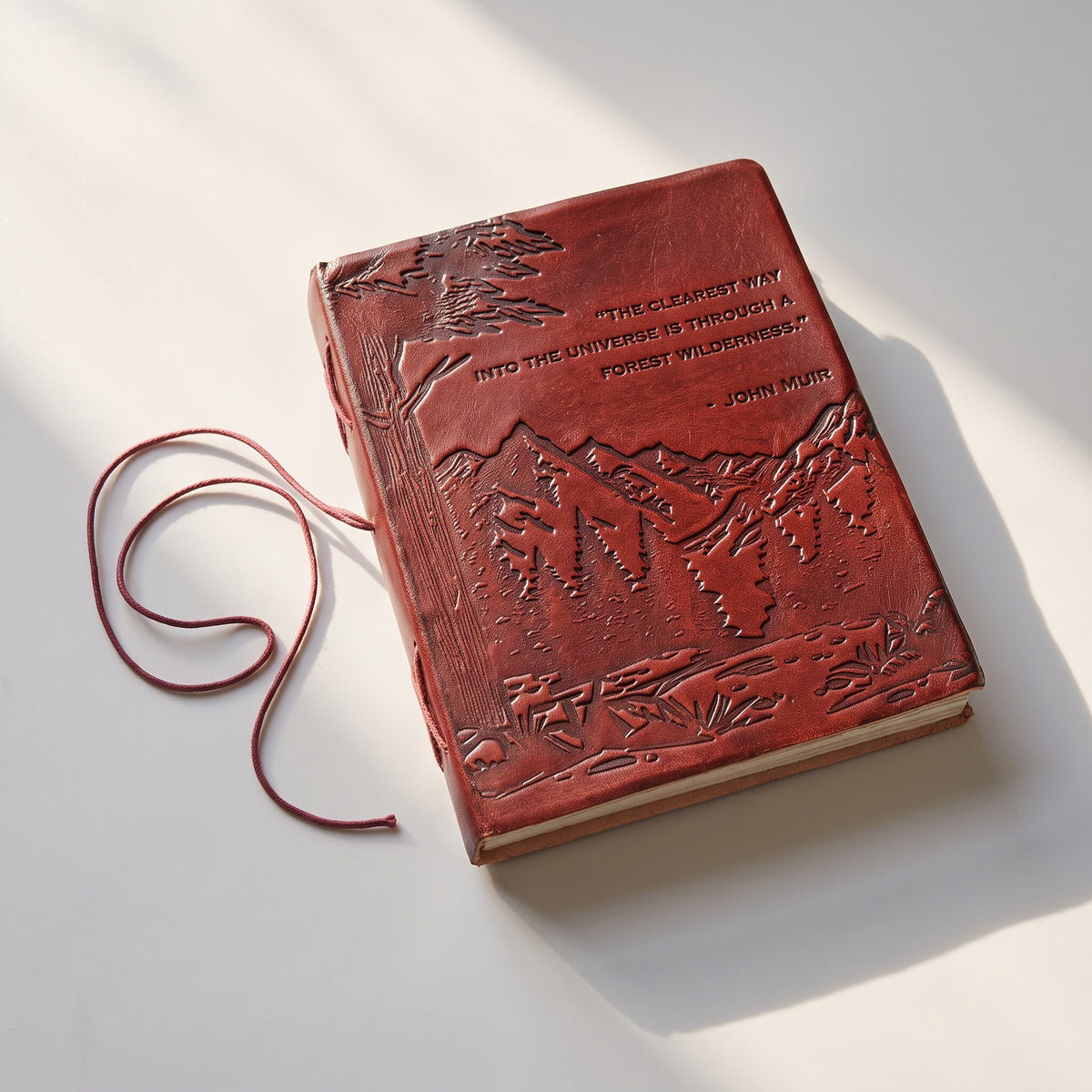 A red leather journal with embossed mountain and tree designs, featuring a John Muir quote on the cover and a matching red ribbon bookmark, photographed on a white surface with soft shadows.
