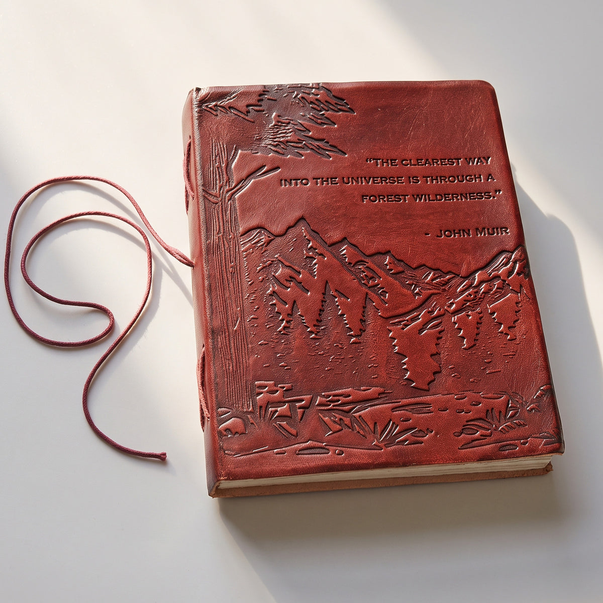 A red leather-bound journal featuring an embossed mountain and forest landscape on the cover, with a red cord bookmark and a John Muir quote embossed on the front, photographed on a white surface with soft shadows.