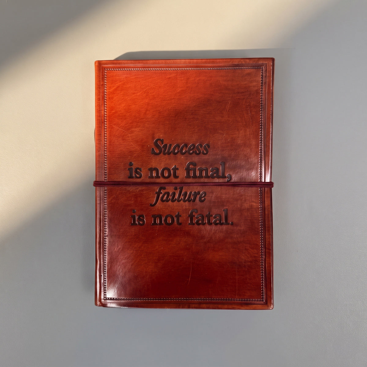 A reddish-brown leather journal with an embossed quote on the cover, 'Success is not final, failure is not fatal.' held in place by a matching leather band, set against a neutral gray background with soft diagonal shadows.