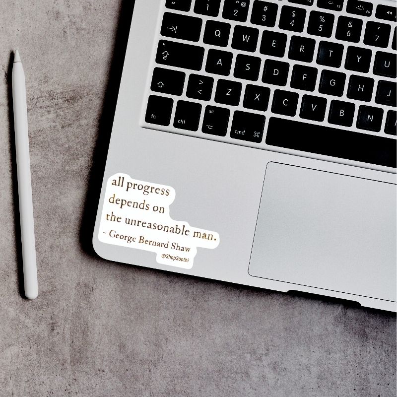 A silver laptop on a gray textured surface with a white stylus beside it. A small sticker on the palm rest reads, 'all progress depends on the unreasonable man.